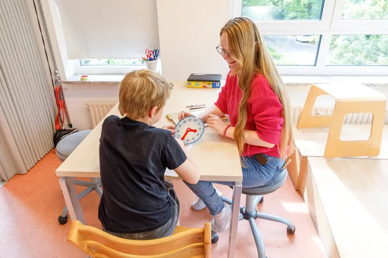Ergotherapeutin lernt mit einem Kind am Tisch die Uhr lesen